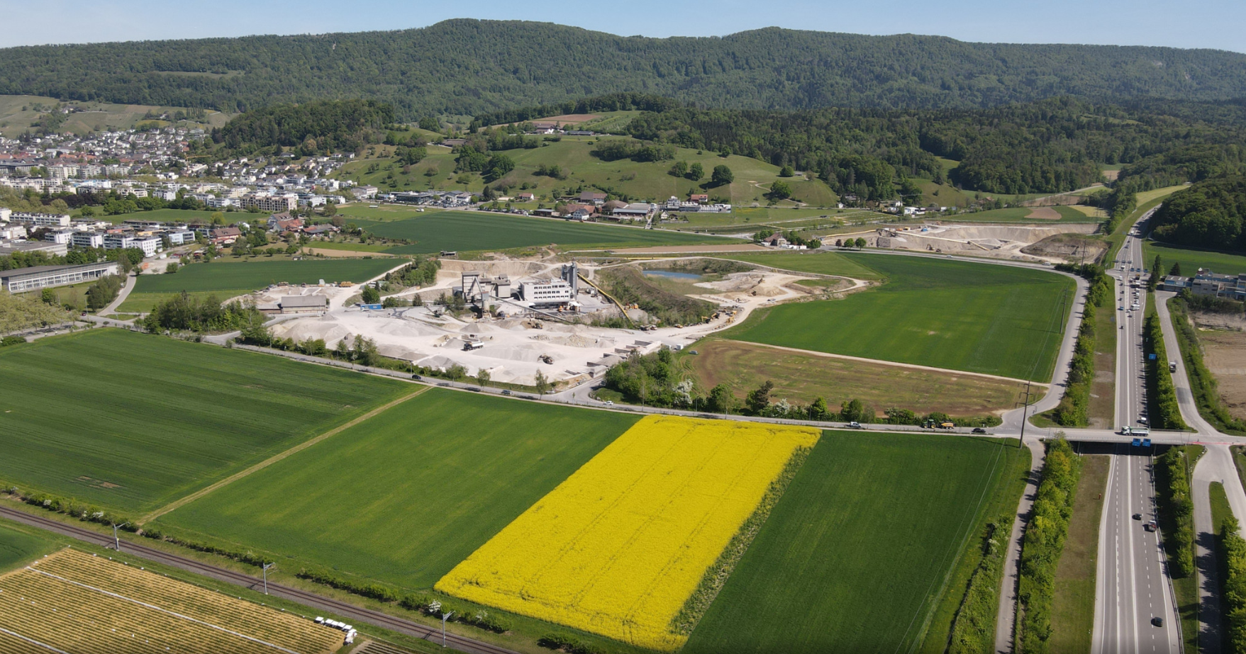 Luftaufnahme einer Landschaft mit gelben Rapsfeldern, grünen Wiesen, einer Industrieanlage und einer Ortschaft vor bewaldeten Hügeln.