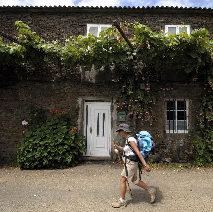 Un pèlerin marche le long du chemin de Saint-Jacques près du village d’O Pino, à 20 km de Saint-Jacques-de-Compostelle, devant une maison en pierre avec des plantes grimpantes.