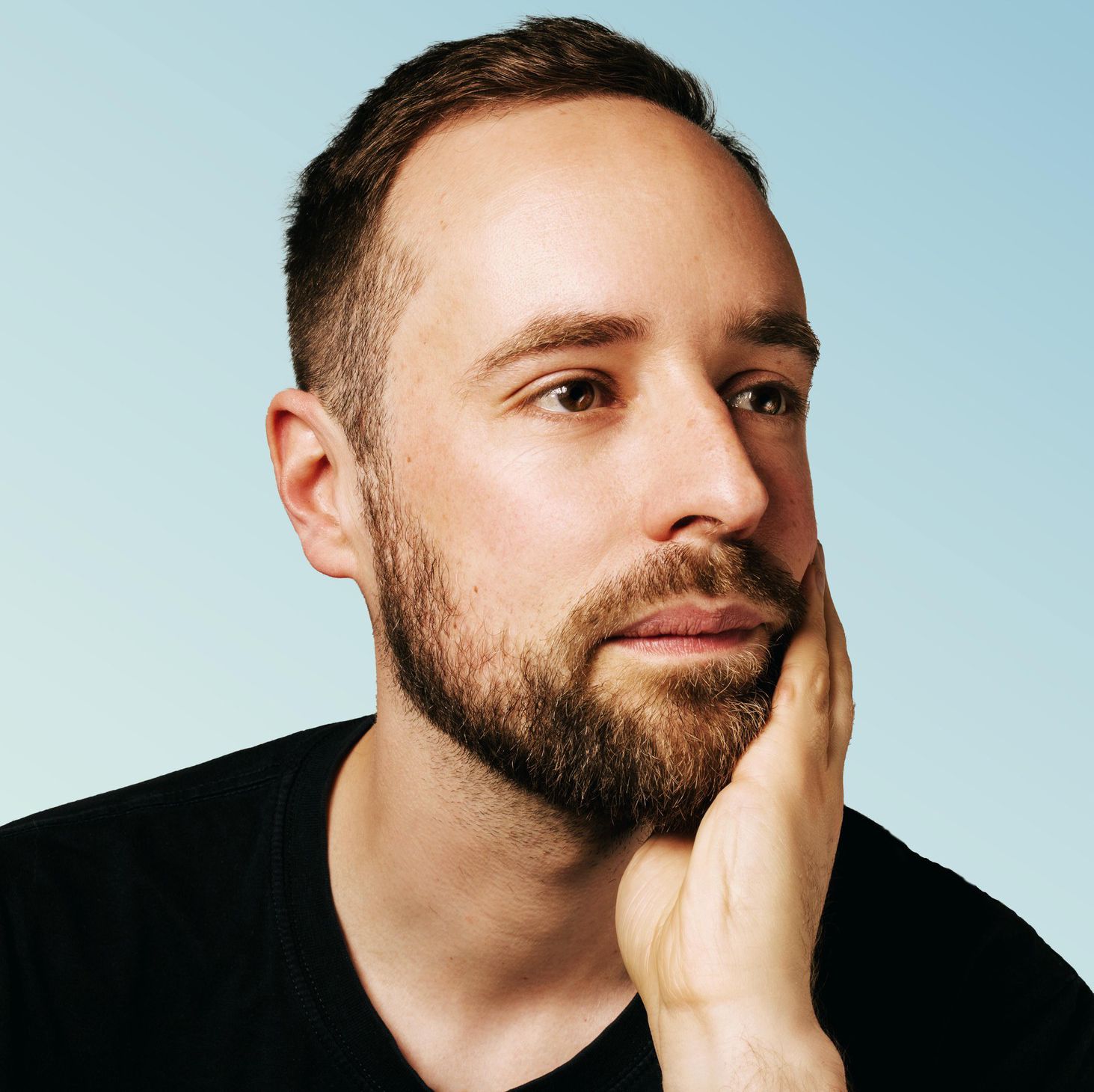 Un homme barbu pose pensivement devant un fond bleu clair.