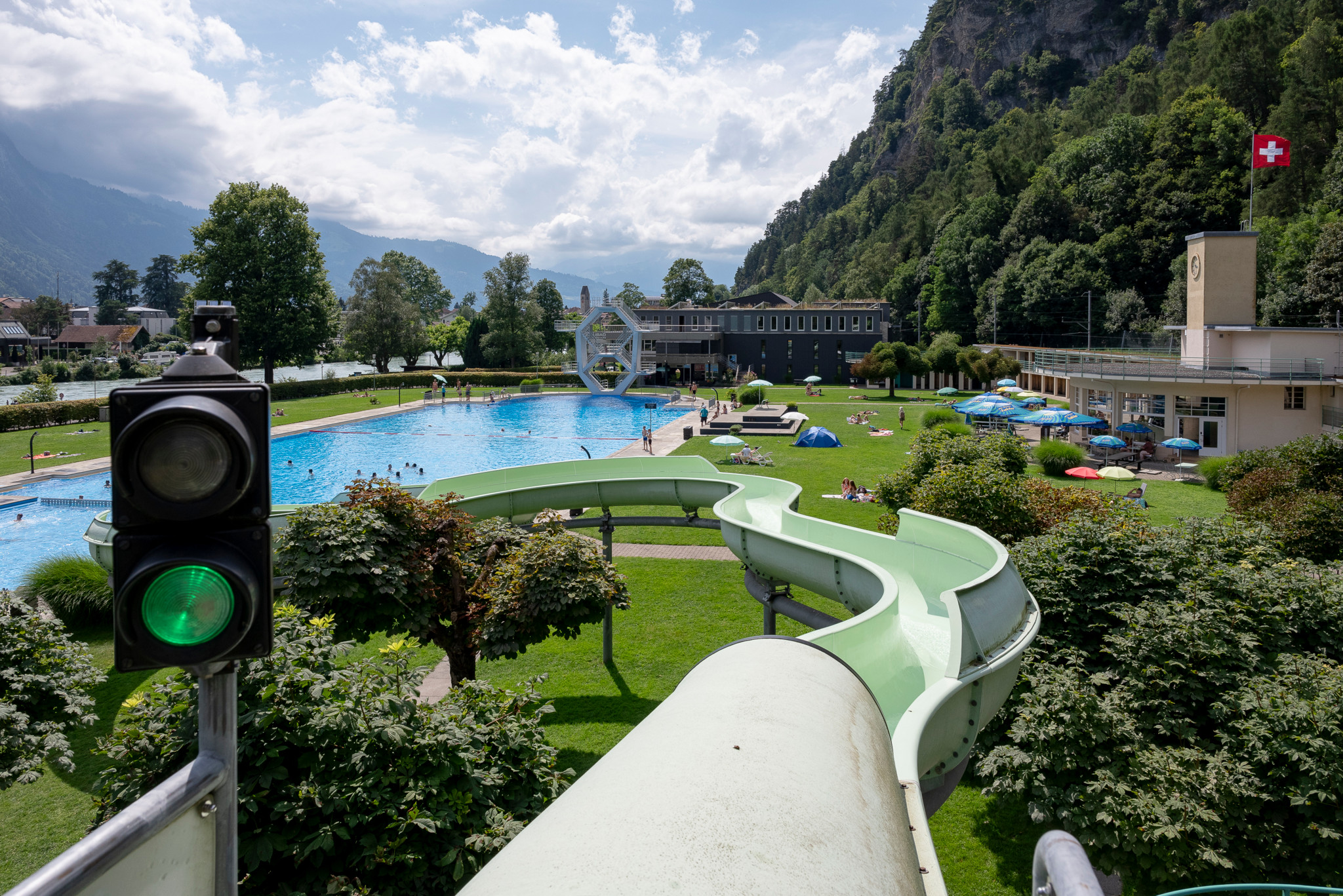 Bödelibad Interlaken mit grüner Wasserrutsche, Sprungturm und Schwimmbecken, umgeben von Bergen und Schweizer Flagge. Bödelibad Interlaken mit grüner Wasserrutsche, Sprungturm und Schwimmbecken, umgeben von Bergen und Schweizer Flagge.
