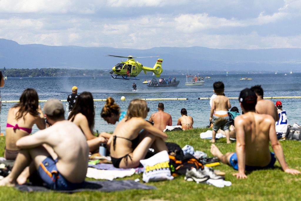 Des personnes profitent d'une journée ensoleillée sur une plage au bord d'un lac, avec un hélicoptère jaune survolant l'eau.