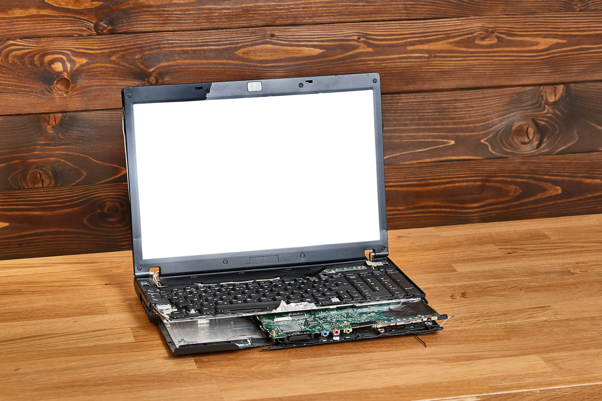 Defekter Laptop mit weissem Bildschirm und freiliegenden Platinen auf einem Holztisch vor Holzwand.