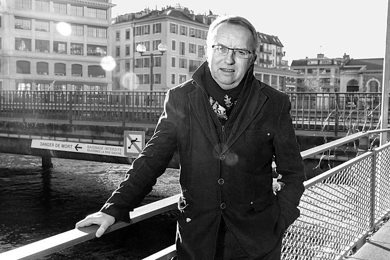 Man in dark coat leaning on a railing near a canal in Geneva, with a 'Danger of Death' sign visible in the background.