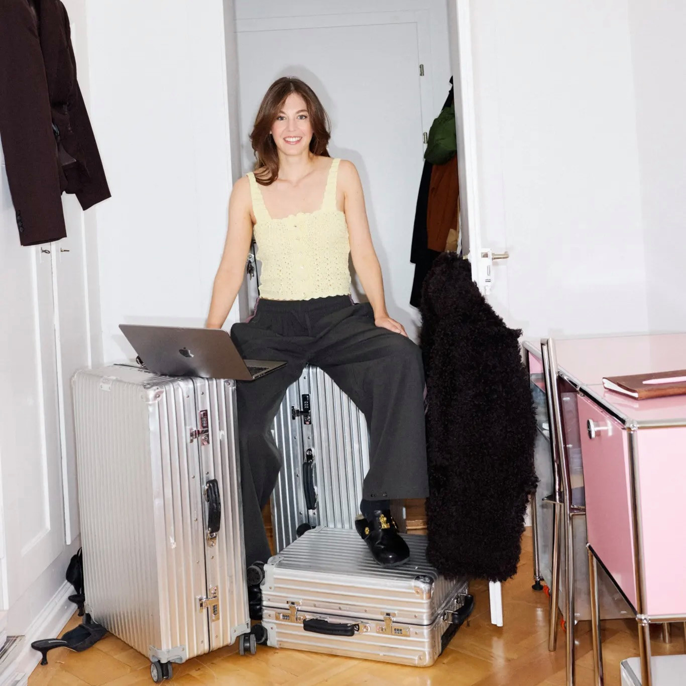 Junge Frau sitzt lächelnd auf Rimowa-Koffern mit Laptop, umgeben von Gepäck und Kleidung in einem hellen Zimmer.
