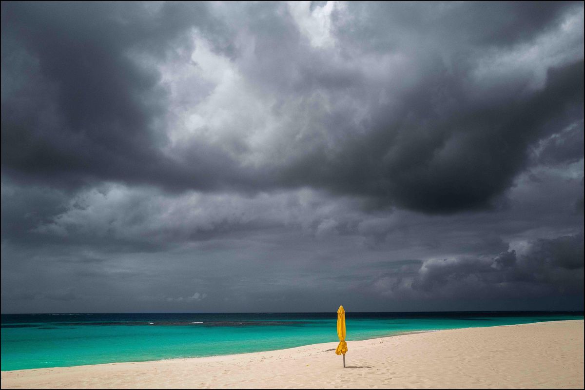 Plage de sable blanc avec un parasol jaune sous un ciel nuageux et menaçant au-dessus de la mer turquoise.