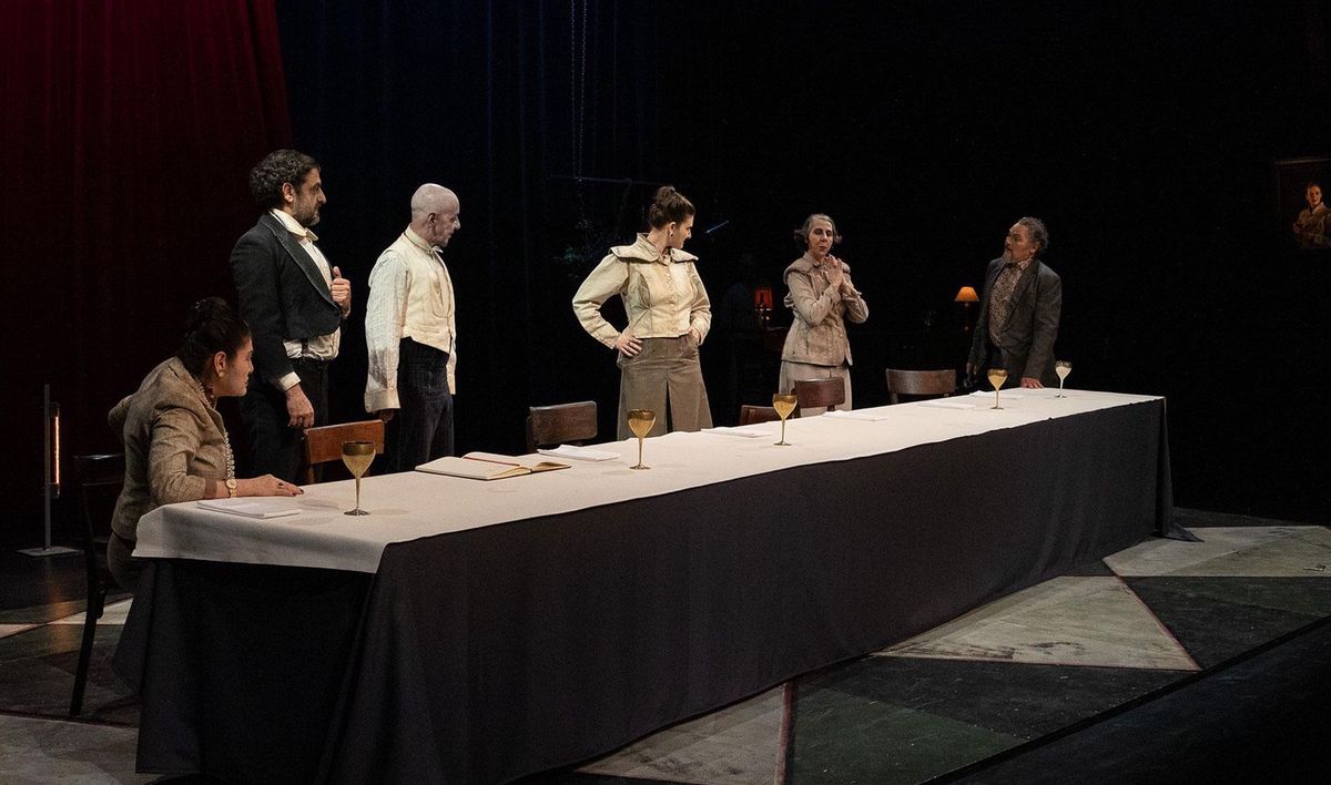 Scène théâtrale sombre avec plusieurs acteurs debout autour d'une table longue avec nappe blanche.