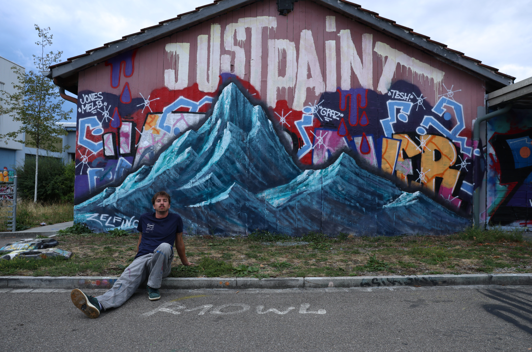 Mann sitzt vor einem bunten Graffiti-Wandgemälde mit Bergen und dem Schriftzug «Just Paint» auf einer Hausfassade.