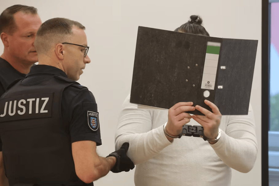 Person in einem Gerichtssaal hält einen Aktenordner vor das Gesicht, während zwei Justizbeamte daneben stehen.