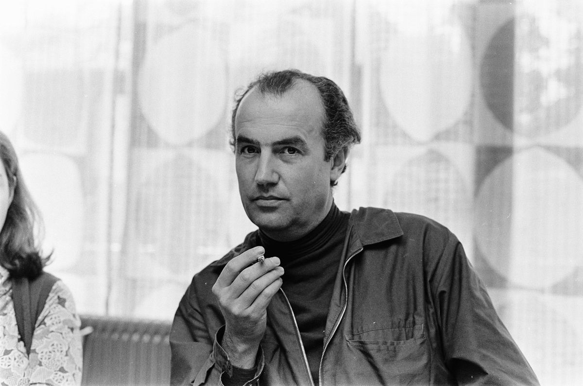 Homme tenant un petit objet dans sa main, portant une veste sombre et un col roulé, assis à l’intérieur devant une fenêtre.