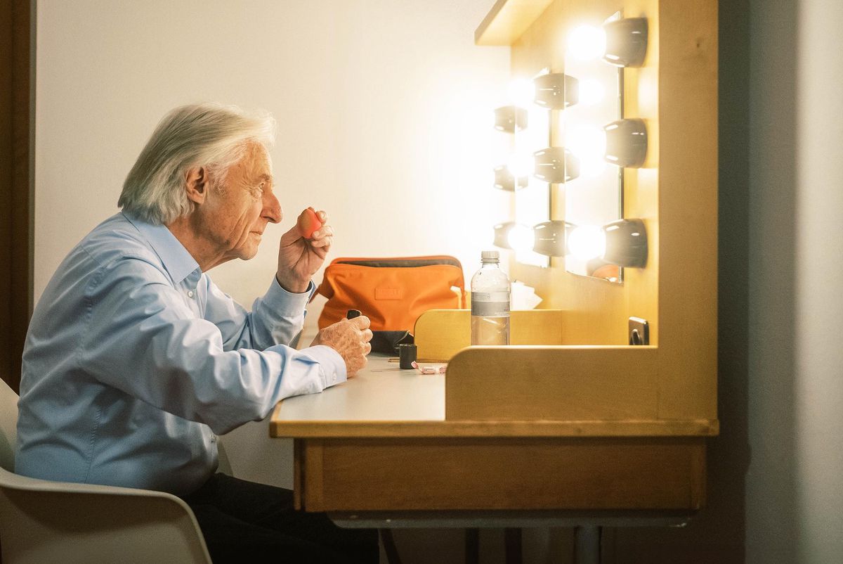 Un homme âgé se prépare devant un miroir éclairé, appliquant du maquillage dans une loge.