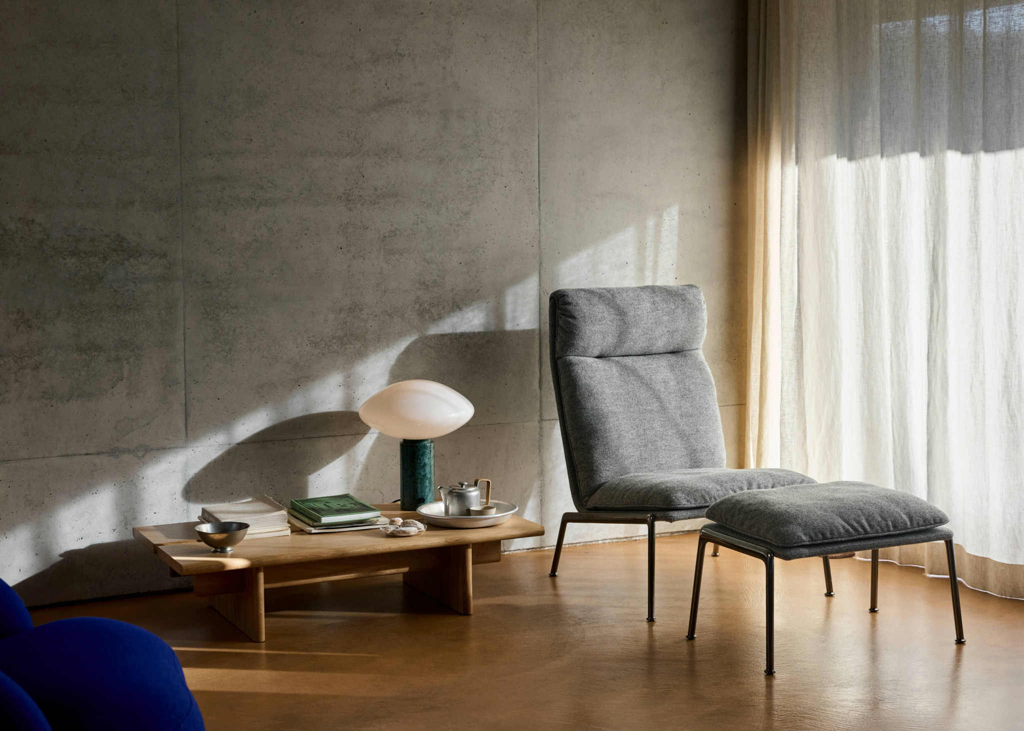 Modernes Wohnzimmer mit grauem Sessel und Hocker, Holzcouchtisch mit Lampe und Büchern, Betonwand und hellen Vorhängen.