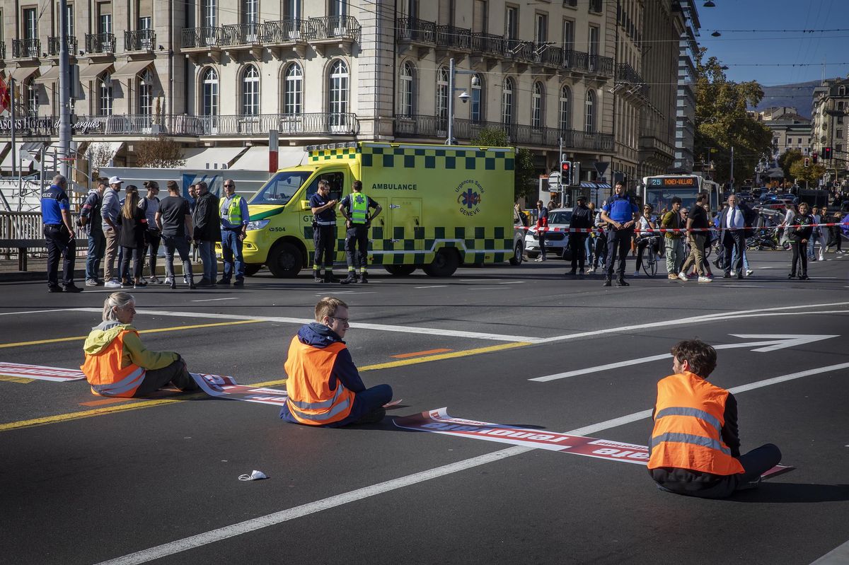 Des activistes de Renovate Switzerland bloquent le Pont du Mont-Blanc à Genève, assis sur la route avec des gilets orange, le 22 octobre 2022. Une ambulance est visible en arrière-plan.