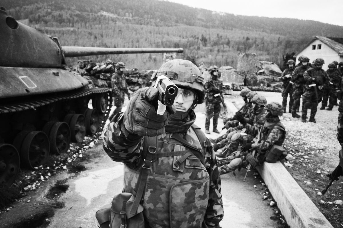 Soldat en uniforme militaire pointant une arme devant un char, entouré de camarades en hiver.