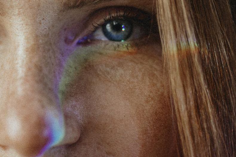 Gros plan d’un visage avec un œil bleu et une lumière arc-en-ciel reflétée sur la peau.