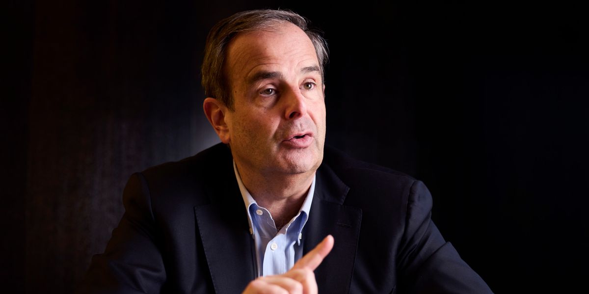 Un homme en costume assis à une table, parlant et faisant un geste de la main avec l'index levé.