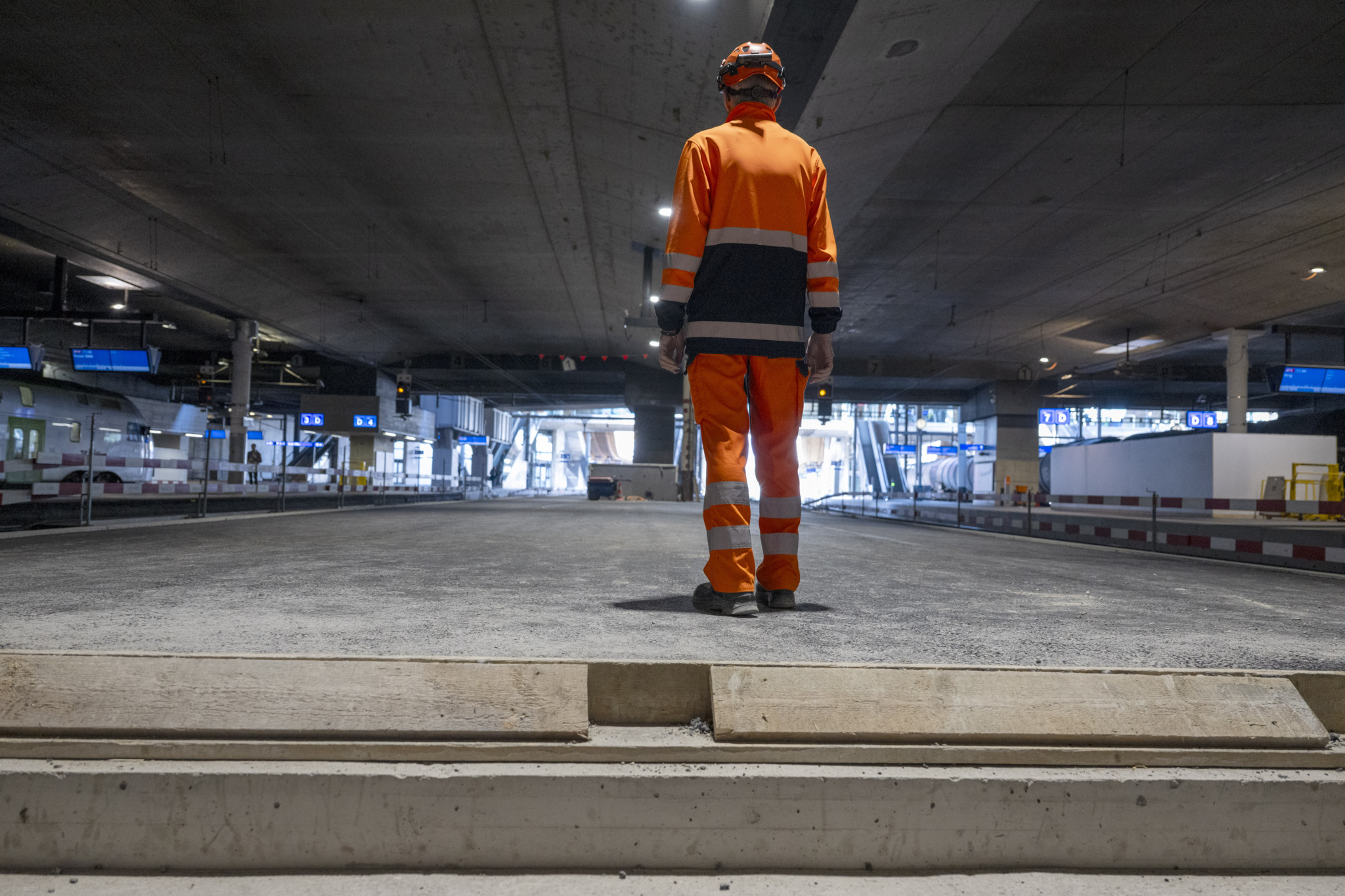 Arbeiter in orangefarbener Schutzkleidung steht auf einer Baustelle im Bahnhof Bern, Perron 3, umgeben von Bauarbeiten und Sicherheitsbarrieren.
