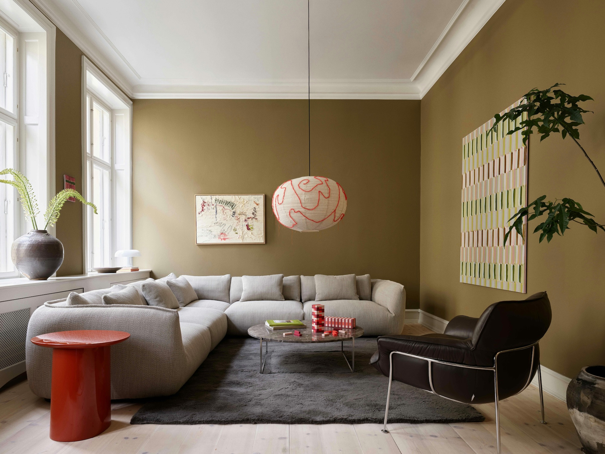 Modernes Wohnzimmer mit olivgrünen Wänden, grauem Ecksofa, rotem Beistelltisch und einer auffälligen rosa Pendelleuchte