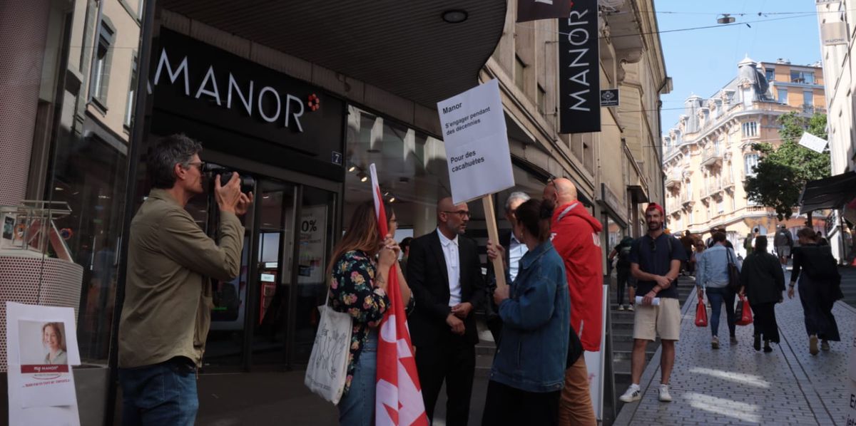 Manifestation devant un magasin Manor en Suisse, avec des gens tenant des pancartes et un photographe prenant des photos.