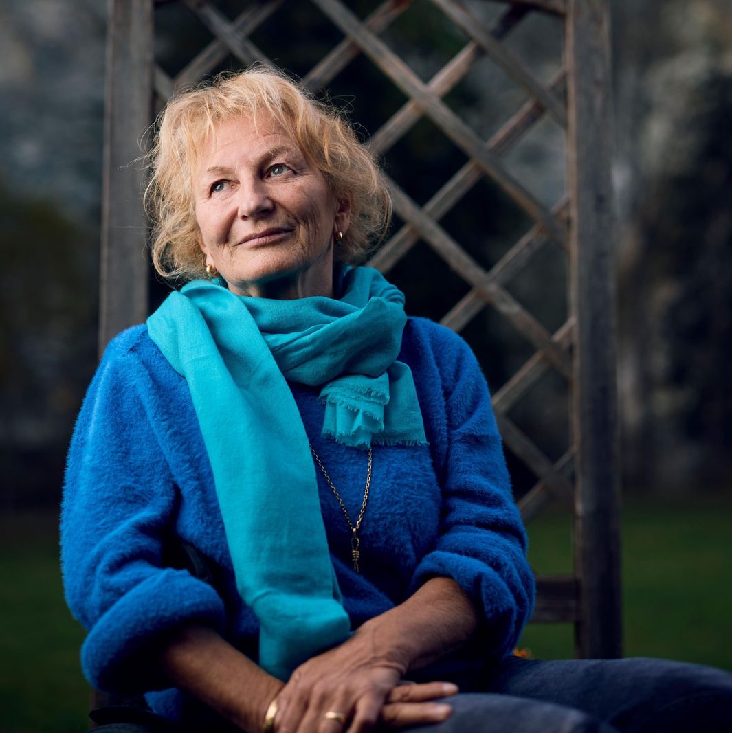 Nicole Niquille assise, portant un pull bleu et une écharpe turquoise, avec un fond naturel à Charmey.