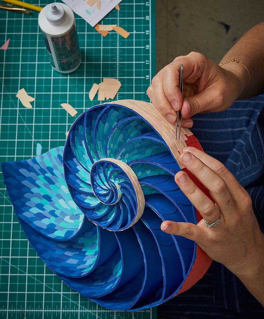 Personne travaillant sur une sculpture en spirale bleue et verte avec des outils d'artisanat et de la colle sur une table de travail.