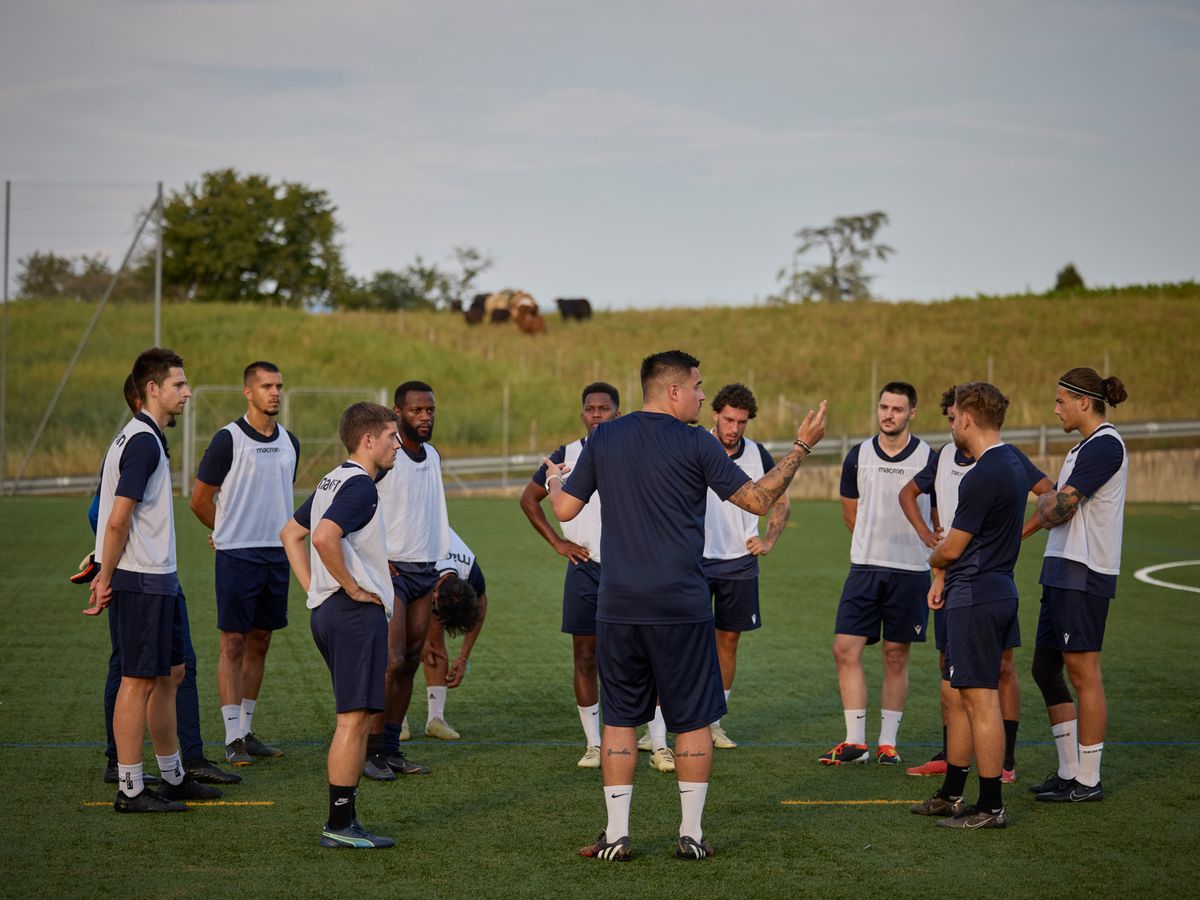Les joueurs du Signal FC Bernex-Confignon en tenue d'entraînement écoutent les instructions de leur entraîneur sur un terrain de football, préparant le match contre le FC Servette-Genève pour le premier tour de la Coupe Suisse.