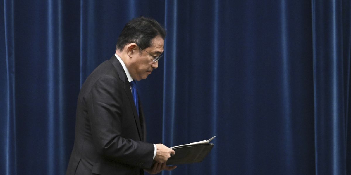 Ein Geschäftsmann in einem schwarzen Anzug und einer blauen Krawatte hält ein Dokument in der Hand und geht vor einem blauen Vorhang vorbei.