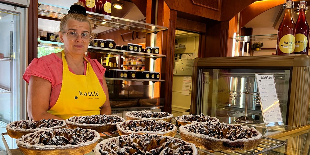 Une femme portant un tablier jaune se tient derrière un présentoir de pâtisseries, avec plusieurs tartes enneigées de sucre en poudre devant elle dans une boulangerie.