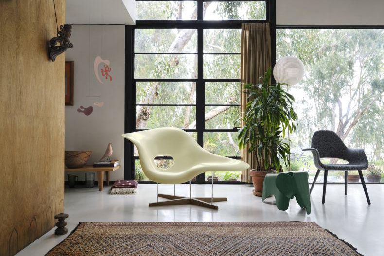 Salon moderne avec de grandes fenêtres laissant entrer la lumière naturelle, fauteuils design, plantes et tapis coloré.