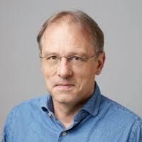 Homme portant une veste bleue, une chemise assortie et un jean, les mains dans les poches, sur un fond gris.