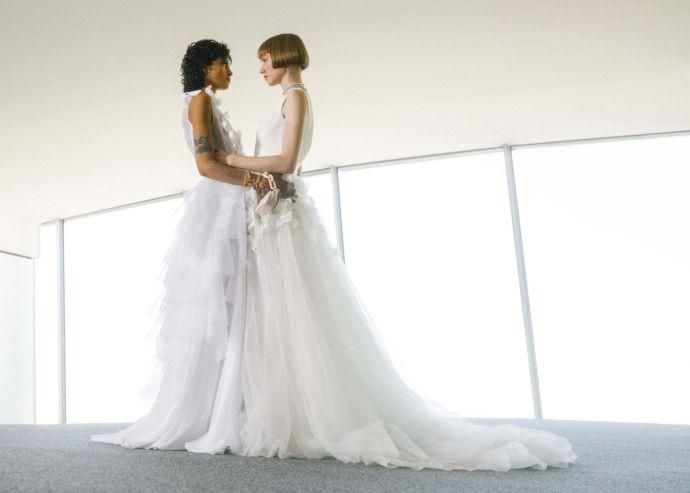 Deux femmes en robes de mariée, face à face, dans une pièce lumineuse avec de grandes fenêtres.