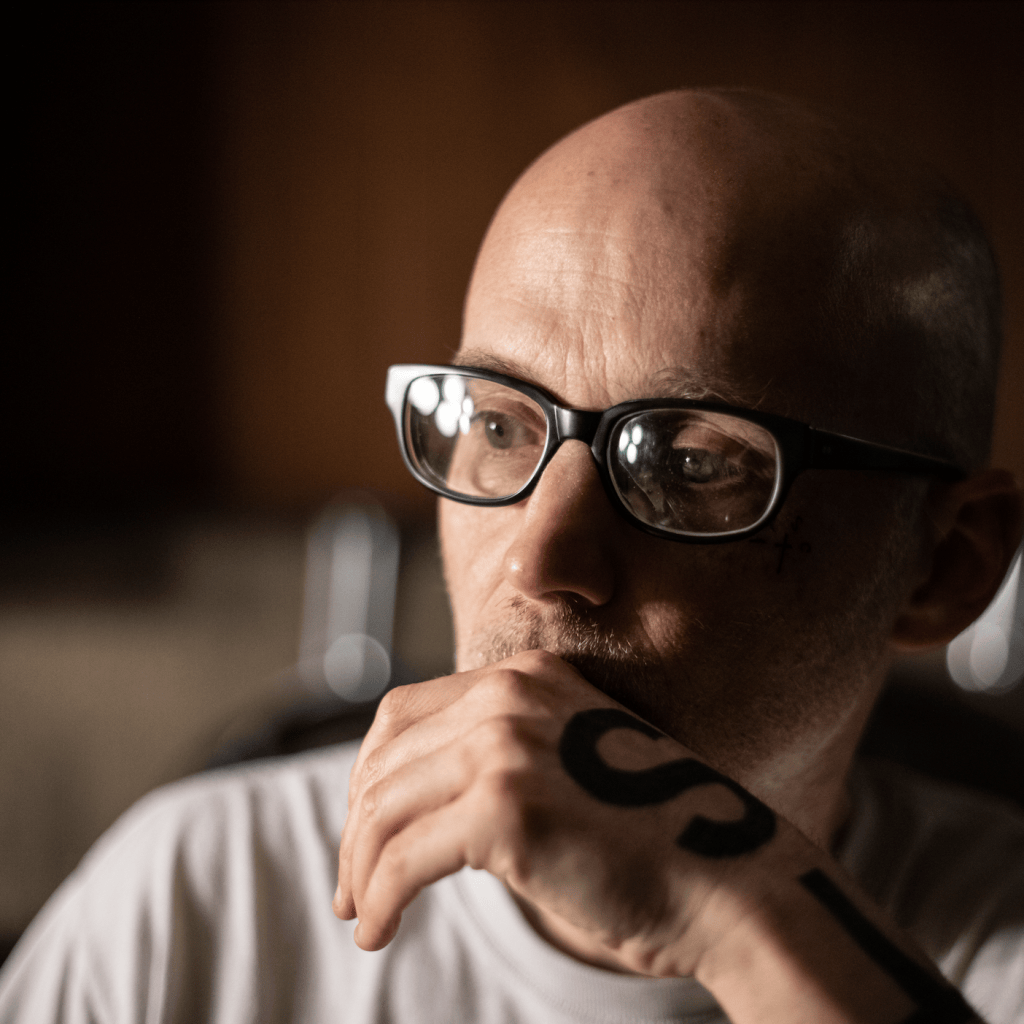 Homme chauve portant des lunettes, la main droite portant un tatouage visible sur sa bouche, regardant pensivement.