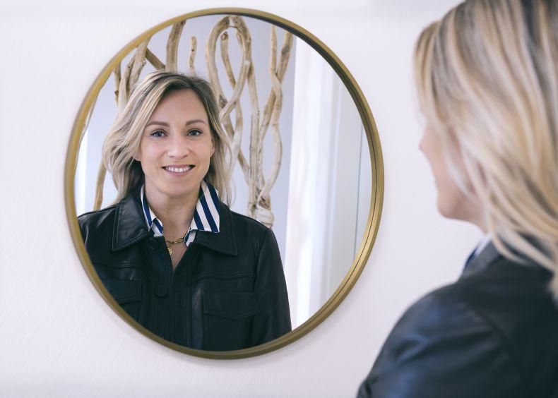 Une femme blonde se reflète dans un miroir rond, portant une veste noire sur une chemise à rayures bleues et blanches.