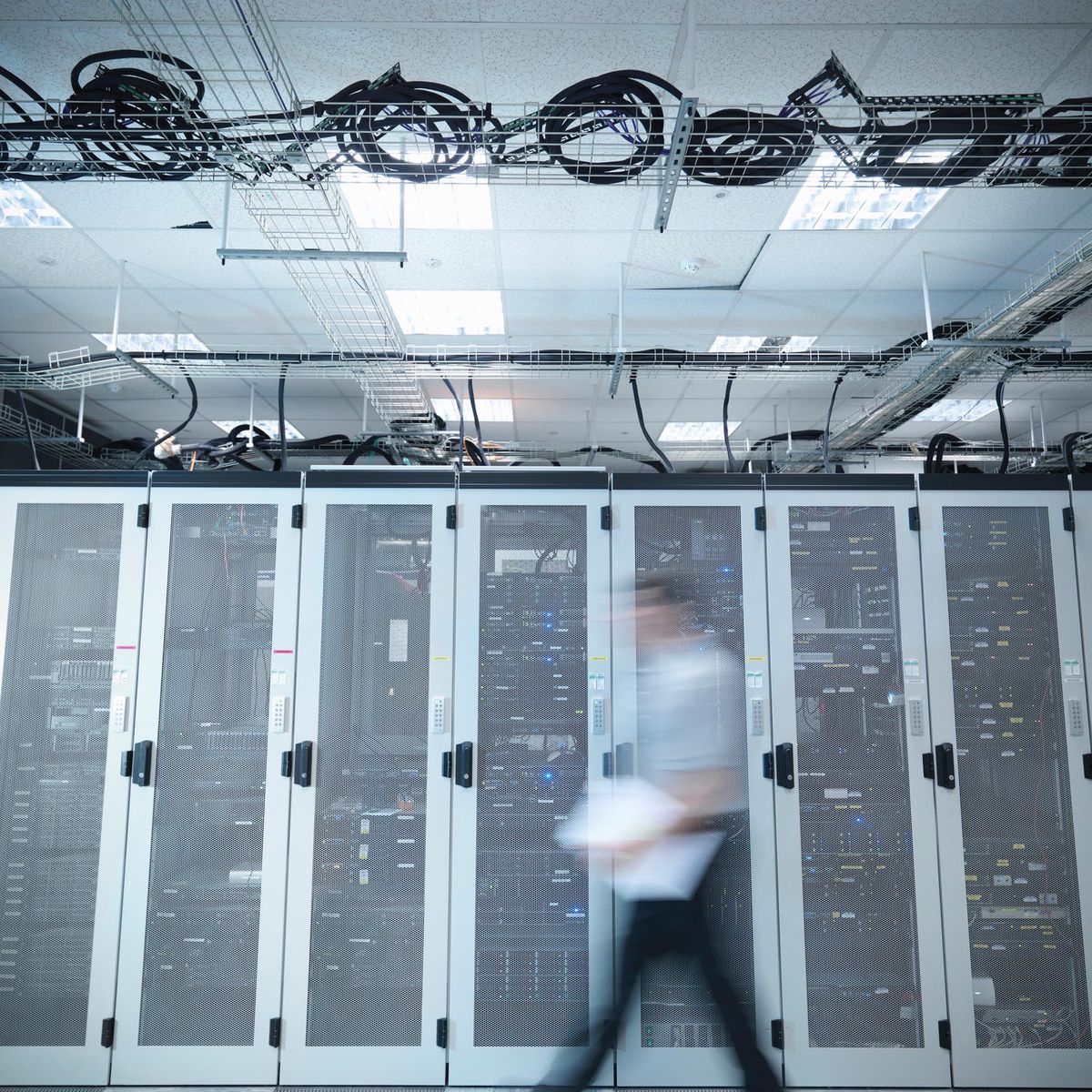 Une personne floue marche devant une rangée de serveurs dans un centre de données, avec câbles visibles au plafond.
