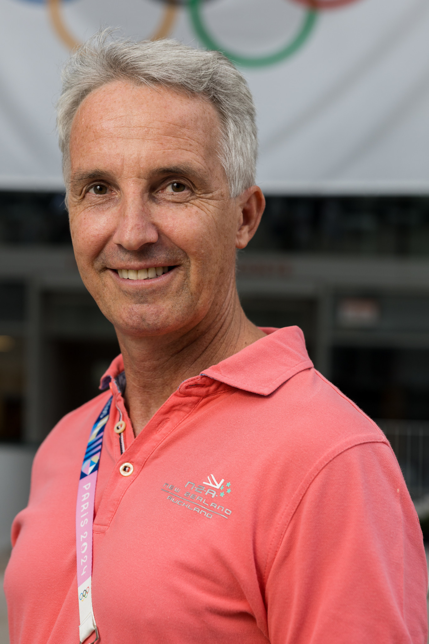 Jean-Laurent Bourquin portant un polo rose, en arrière-plan des anneaux olympiques, lors des JO de Paris 2024 à Roland-Garros.