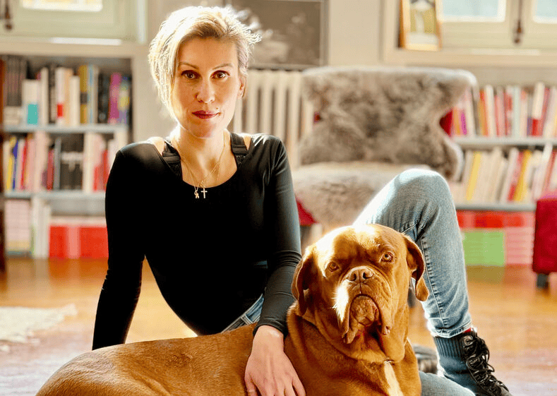 Femme assise avec un chien Dogue de Bordeaux dans un salon lumineux, entourée d’étagères remplies de livres.