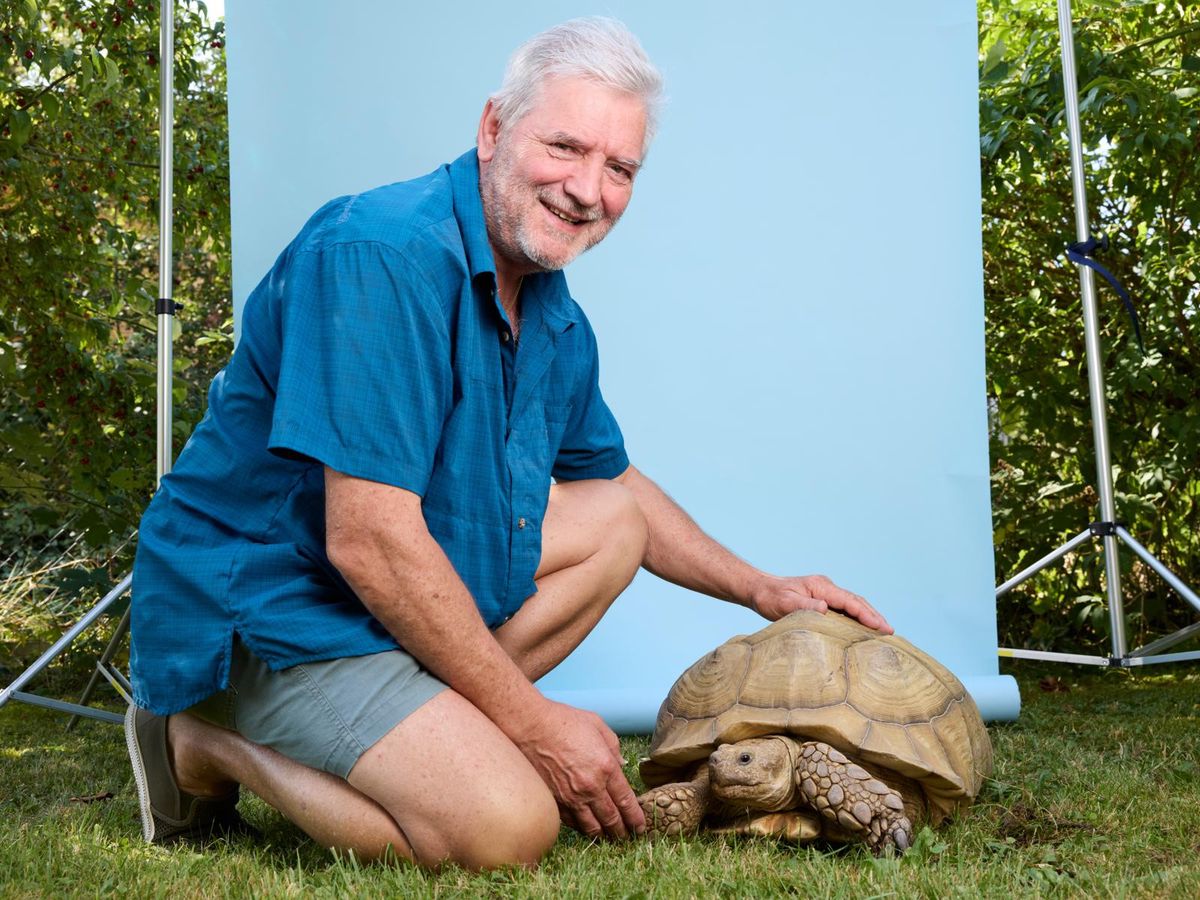 Un homme souriant en chemise bleue et en short gris s'agenouille sur l'herbe à côté d'une grande tortue, posant devant un fond bleu.