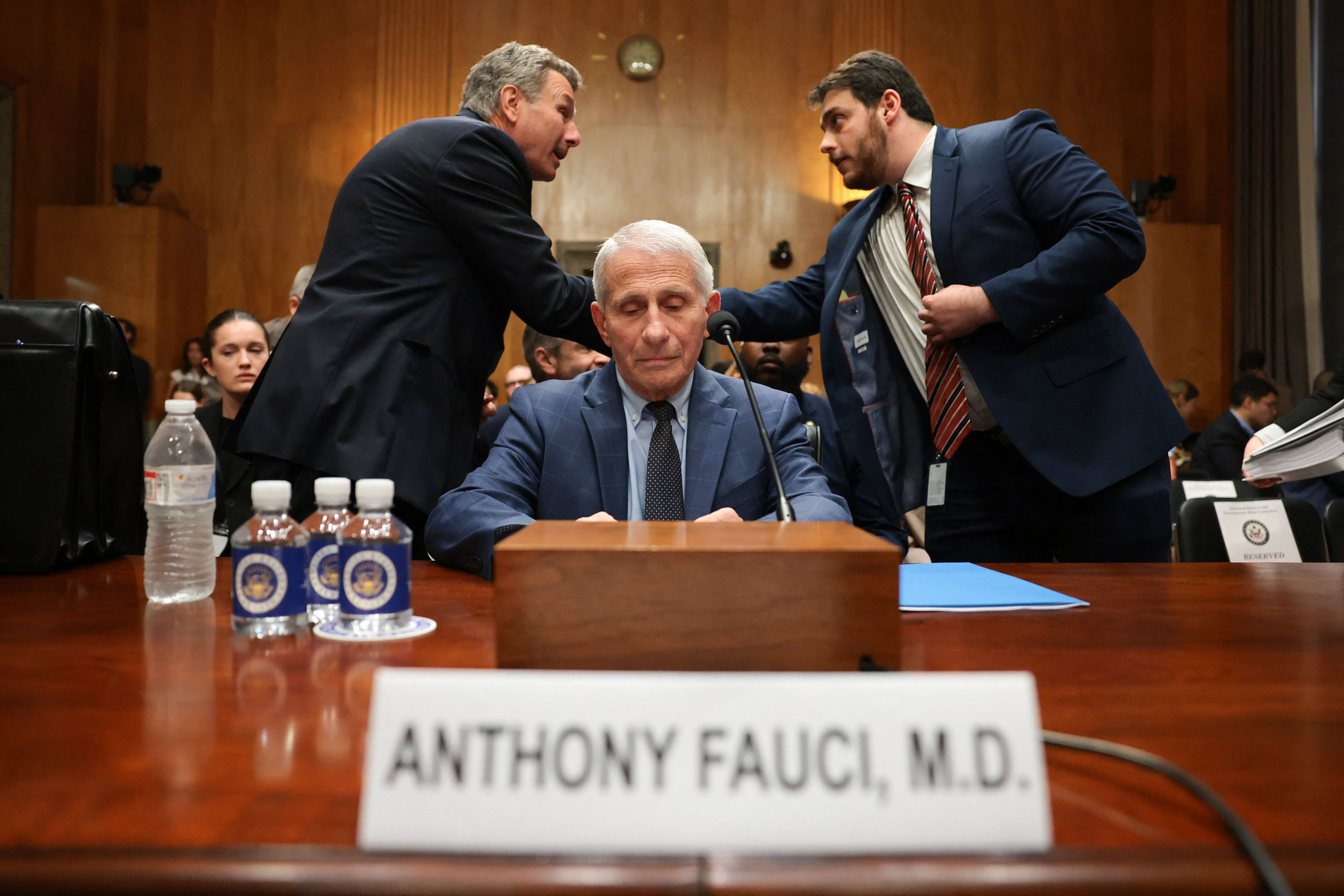 Anthony Fauci, M.D. sitzt vor dem Senatsausschuss in Washington, D.C., während zwei Männer hinter ihm sprechen. Namensschild im Vordergrund.