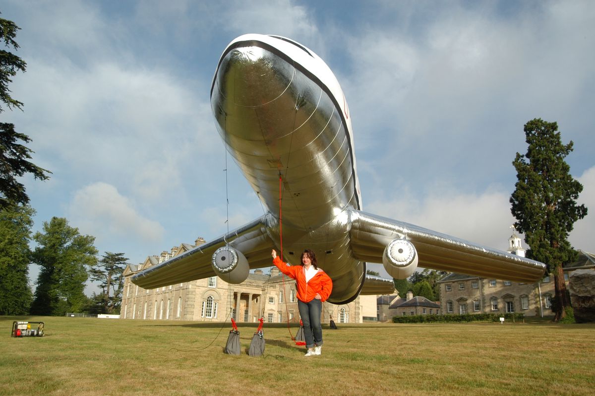 Un grand ballon gonflable en forme d'avion est attaché au sol sur une pelouse devant un bâtiment historique anglais. L'artiste Aleksandra Mir, en blouson orange, tient les câbles du ballon.
