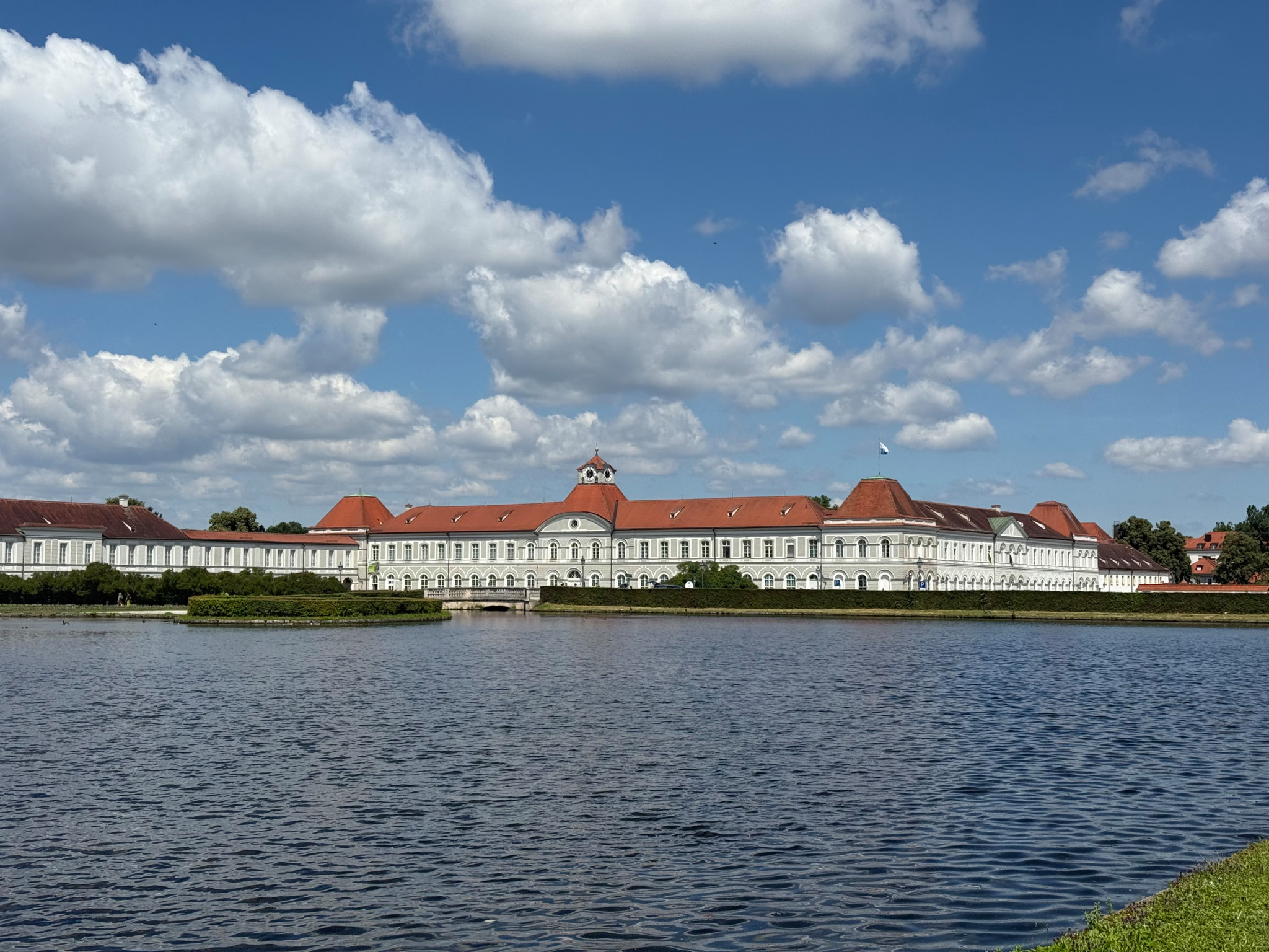 Weitläufiges barockes Schloss mit rotem Dach und weisser Fassade, spiegelt sich in einem ruhigen See unter blauem Sommerhimmel.