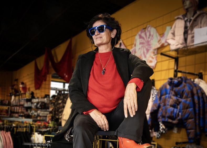 Personne portant des lunettes de soleil bleues et un pull rouge, assise dans un magasin de vêtements. Fonds de manteaux et chemises attachés au mur.