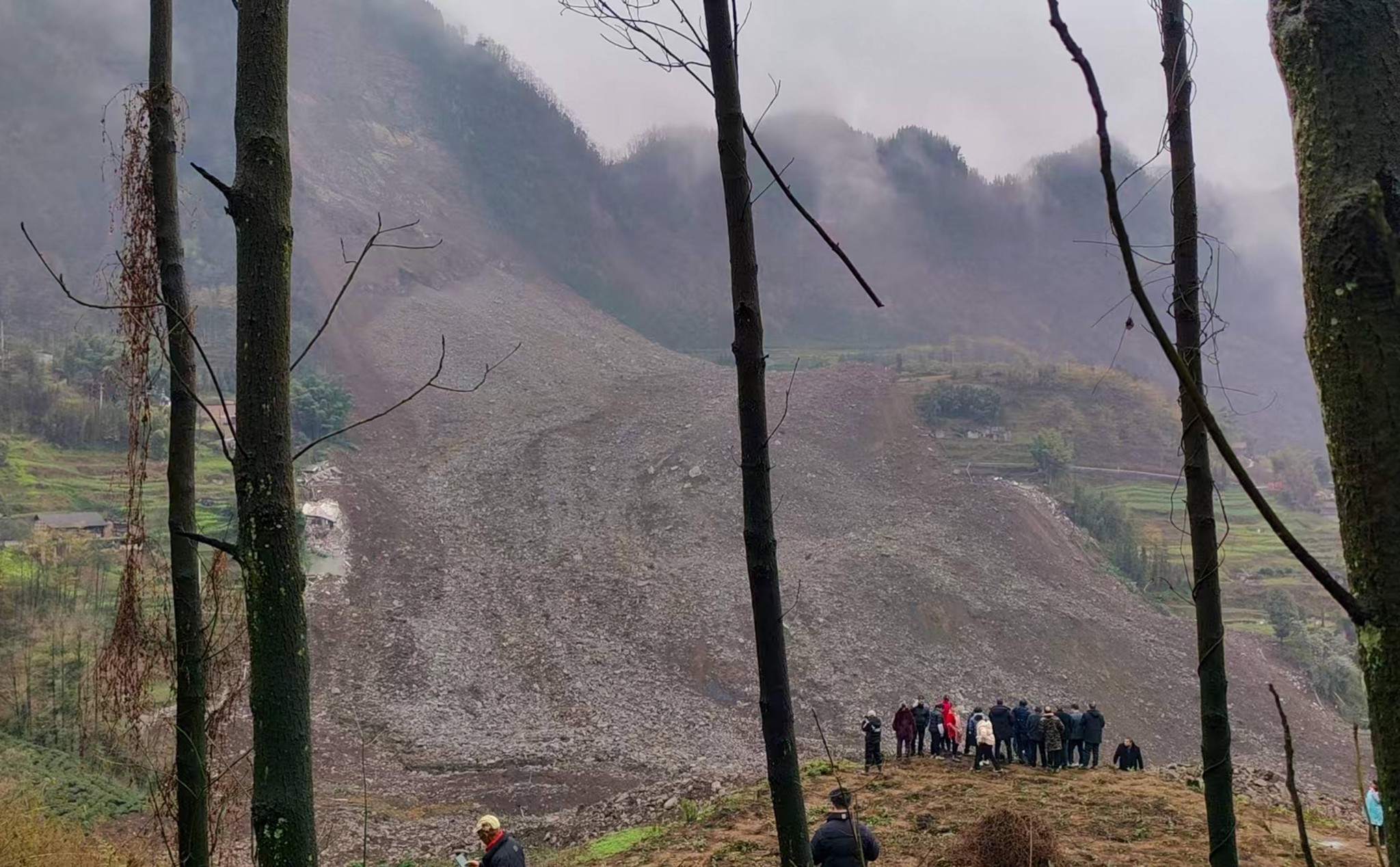 Landschaft mit Erdrutsch in Jinping Village, Junlian County, Yibin, Sichuan, China am 8. Februar 2025. Umgestürzte Erde bedeckt einen Teil der Vegetation, mit einer Gruppe von Menschen im Vordergrund, die die Schäden begutachten.