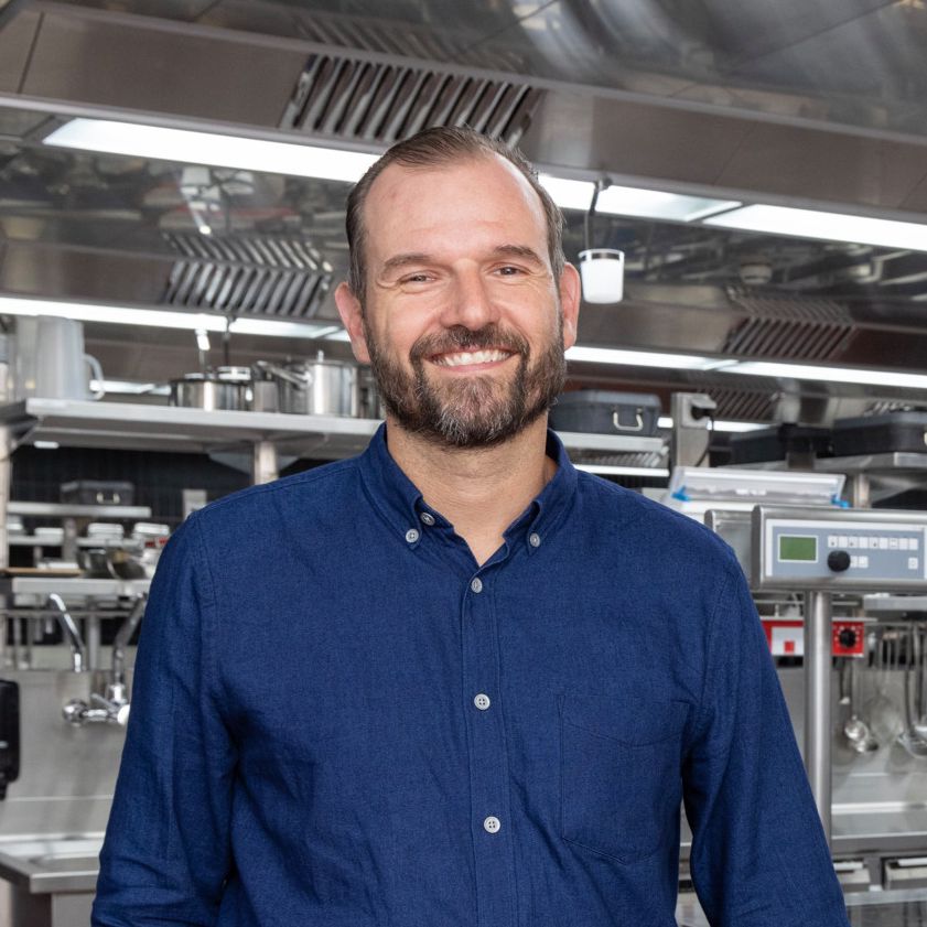 Gastronom Florian Illmer steht lächelnd in einer modernen Grossküche in Zürich, diskutiert über den Fachkräftemangel und die positiven Aspekte des Gastgewerbes.