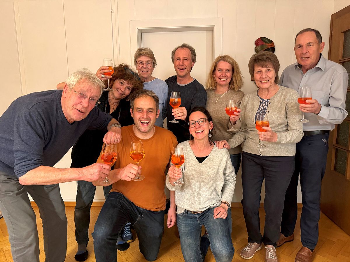 Un groupe de neuf personnes souriantes, tenant des verres de boisson orangée, posant dans une pièce avec un parquet en bois. Certains sont assis, d'autres sont debout.