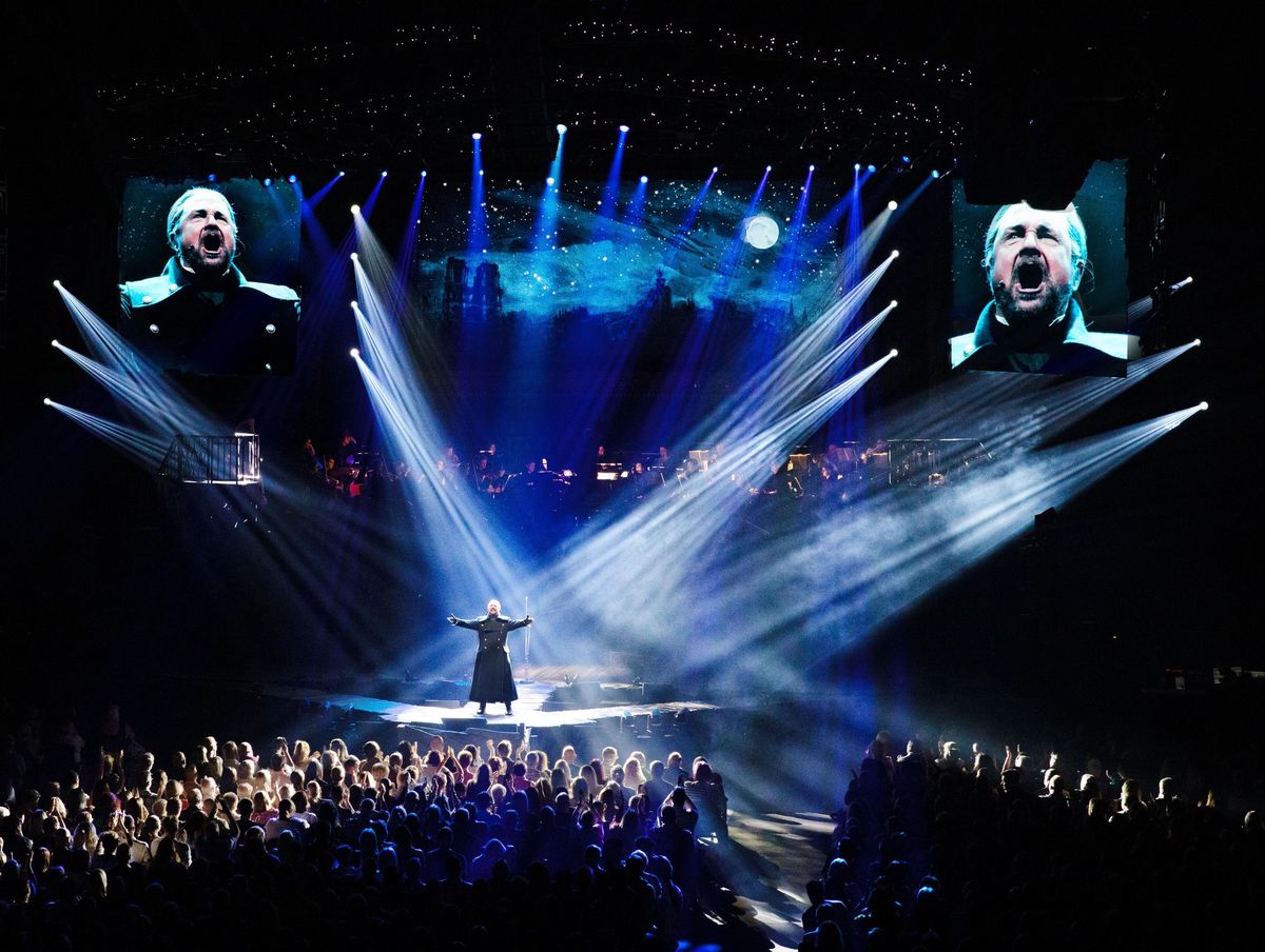 Scène spectaculaire de la comédie musicale 'Les Misérables' avec éclairages dramatiques et deux écrans montrant un acteur exprimant des émotions intenses.