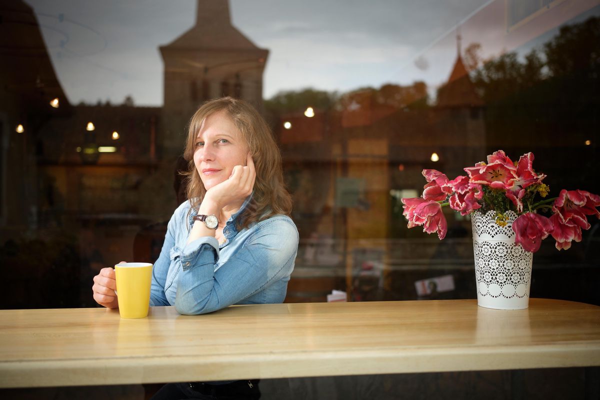 Camille Krafft assise à une table avec une tasse jaune, fleur rose à côté, église en arrière-plan à Romainmotier.